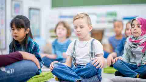 A multi-ethnic group of young school children are indoors in their classroom. They are sitting on pillows and meditating.
